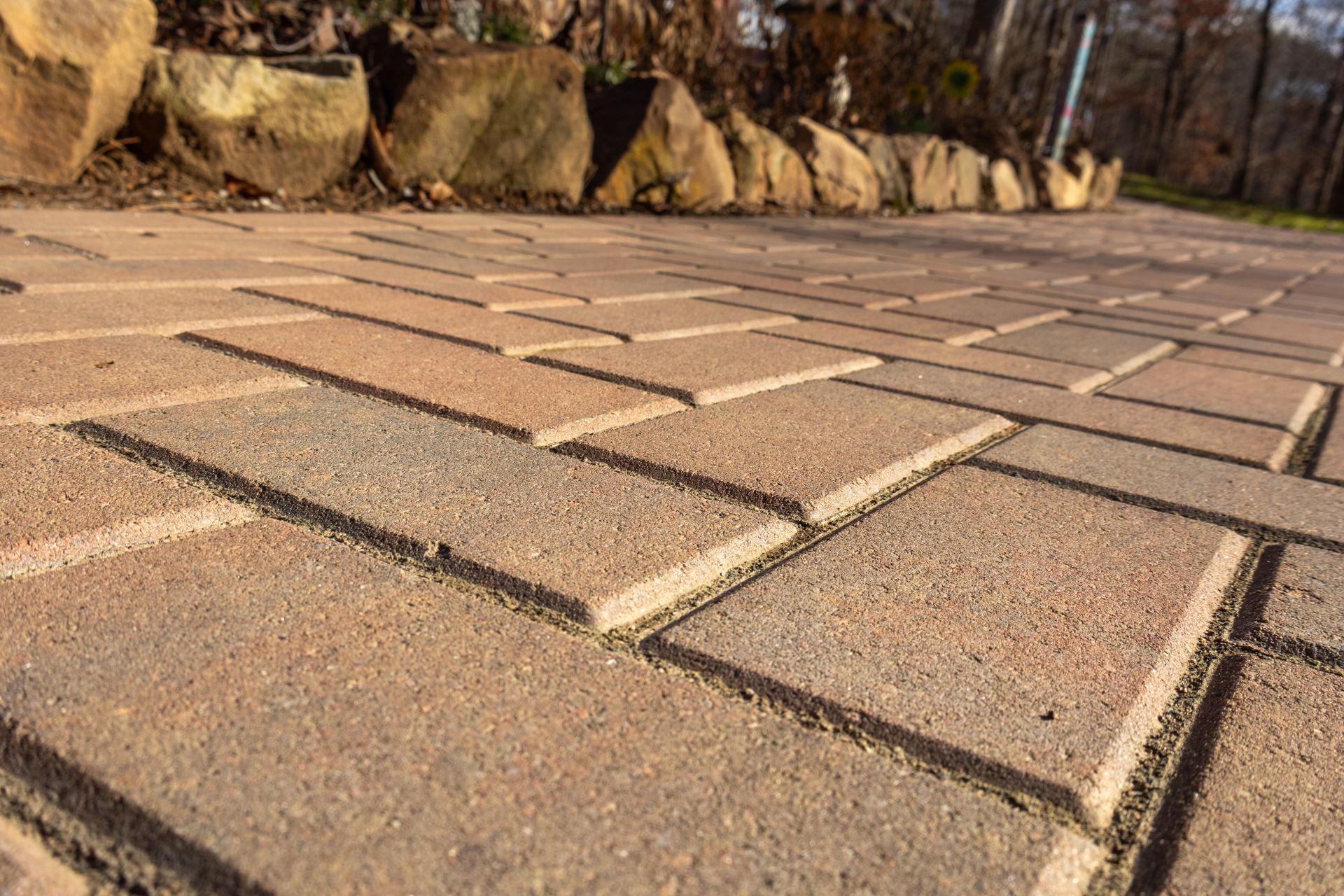 Close-up of brick pathway with light brown bricks, leading to a rock border and trees.