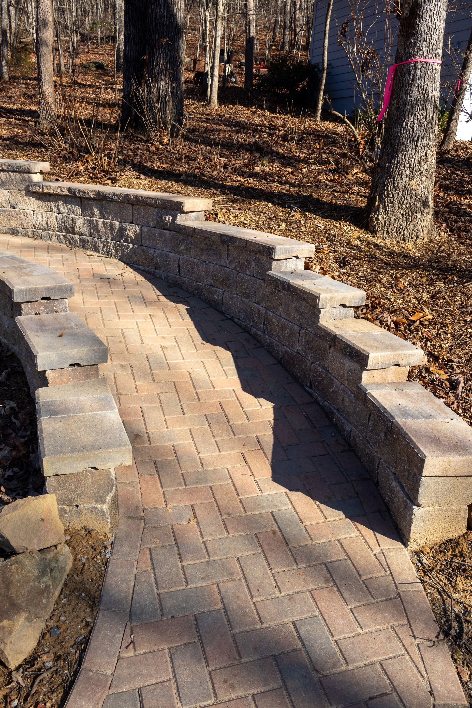 Brick path curves alongside a retaining wall with trees in the background.