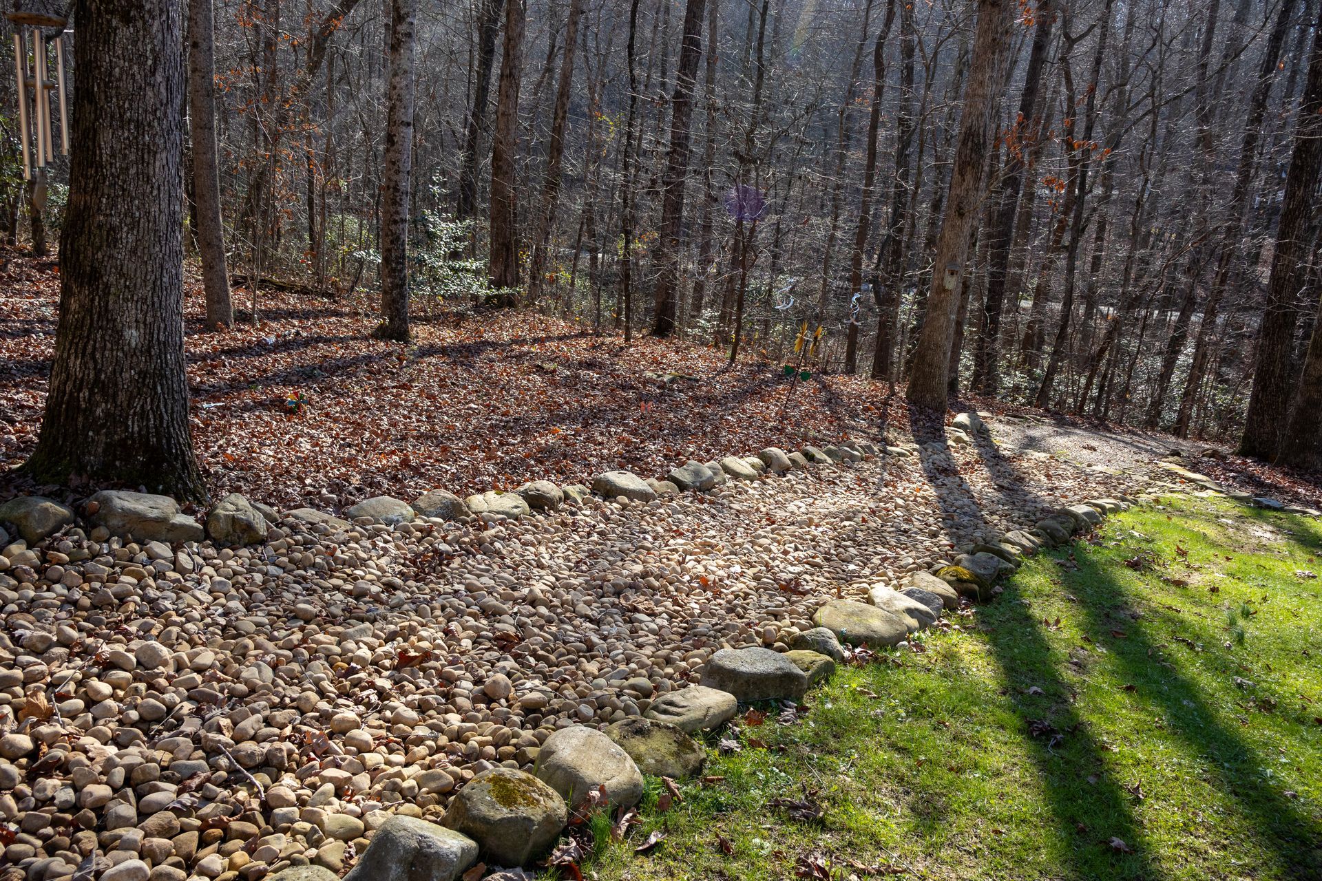 A stone-bordered garden path with pebbles and fallen leaves, next to a grassy area and bare trees.