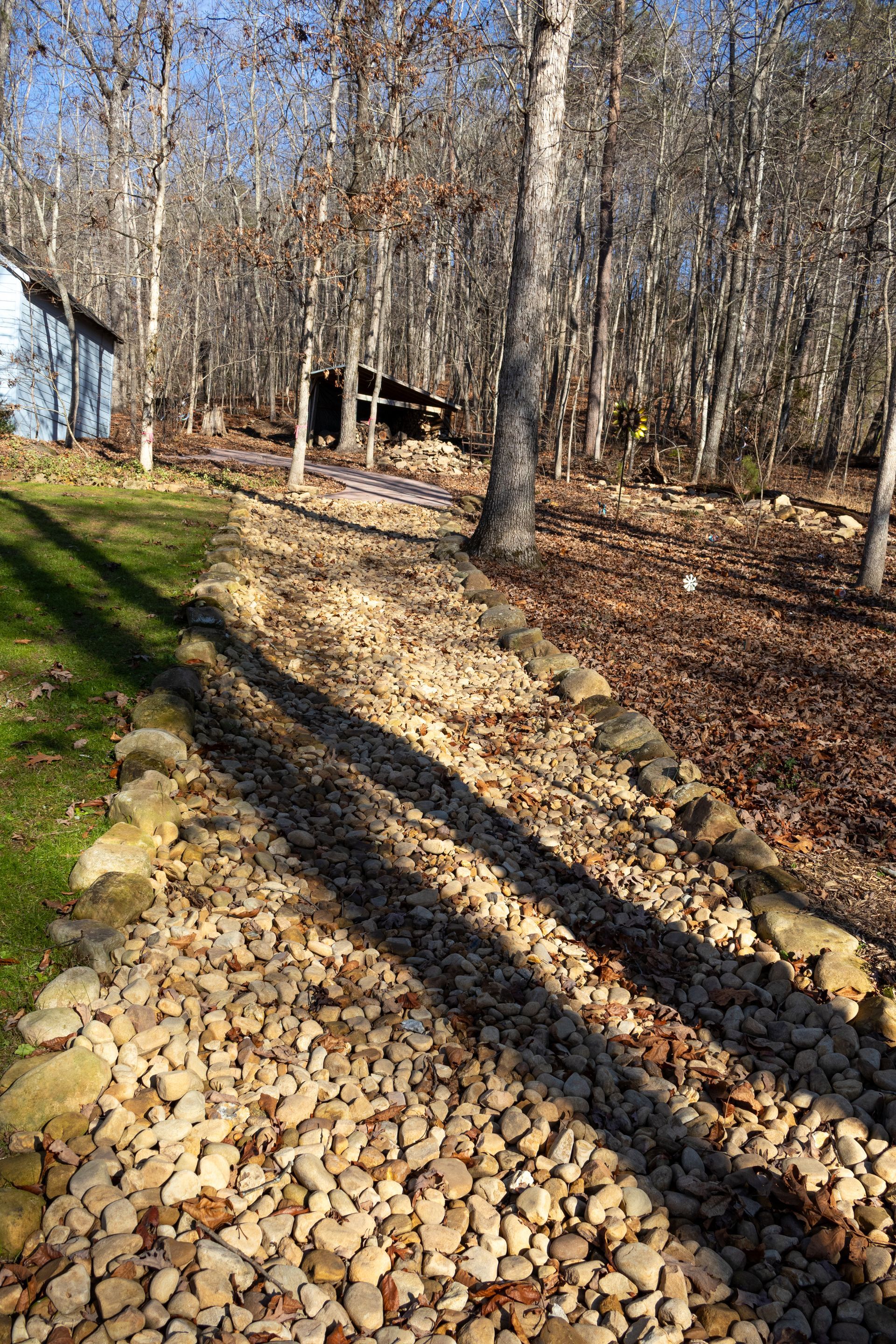 Rocky path with trees, building, and grassy area, casting shadows under a bright blue sky.