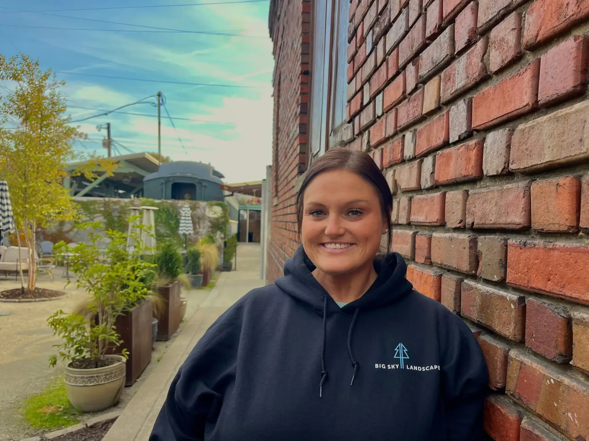 Person smiling beside a brick wall in a courtyard with potted plants and a blue sky