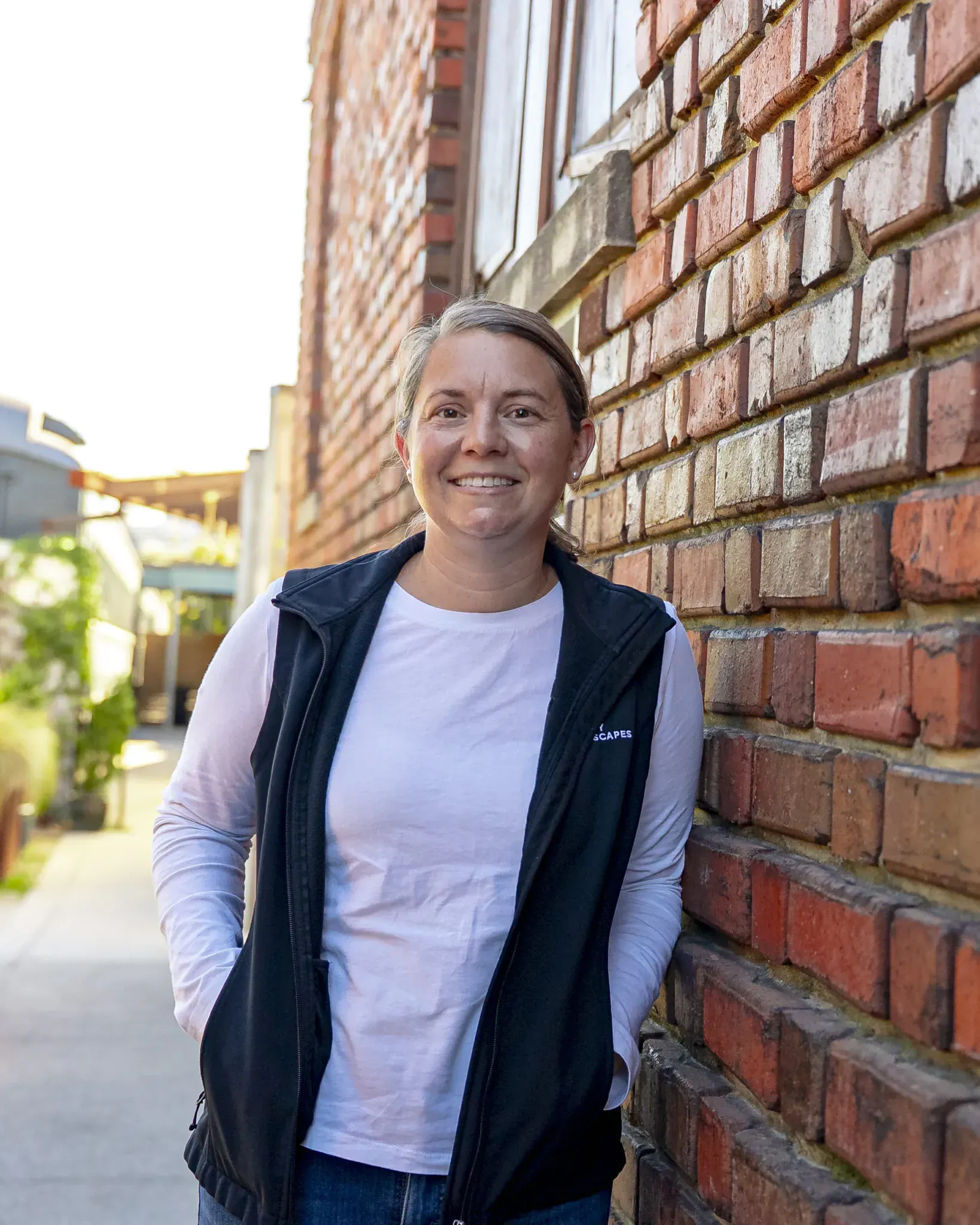 Smiling person leaning against a brick wall in a white shirt and dark vest outdoors.