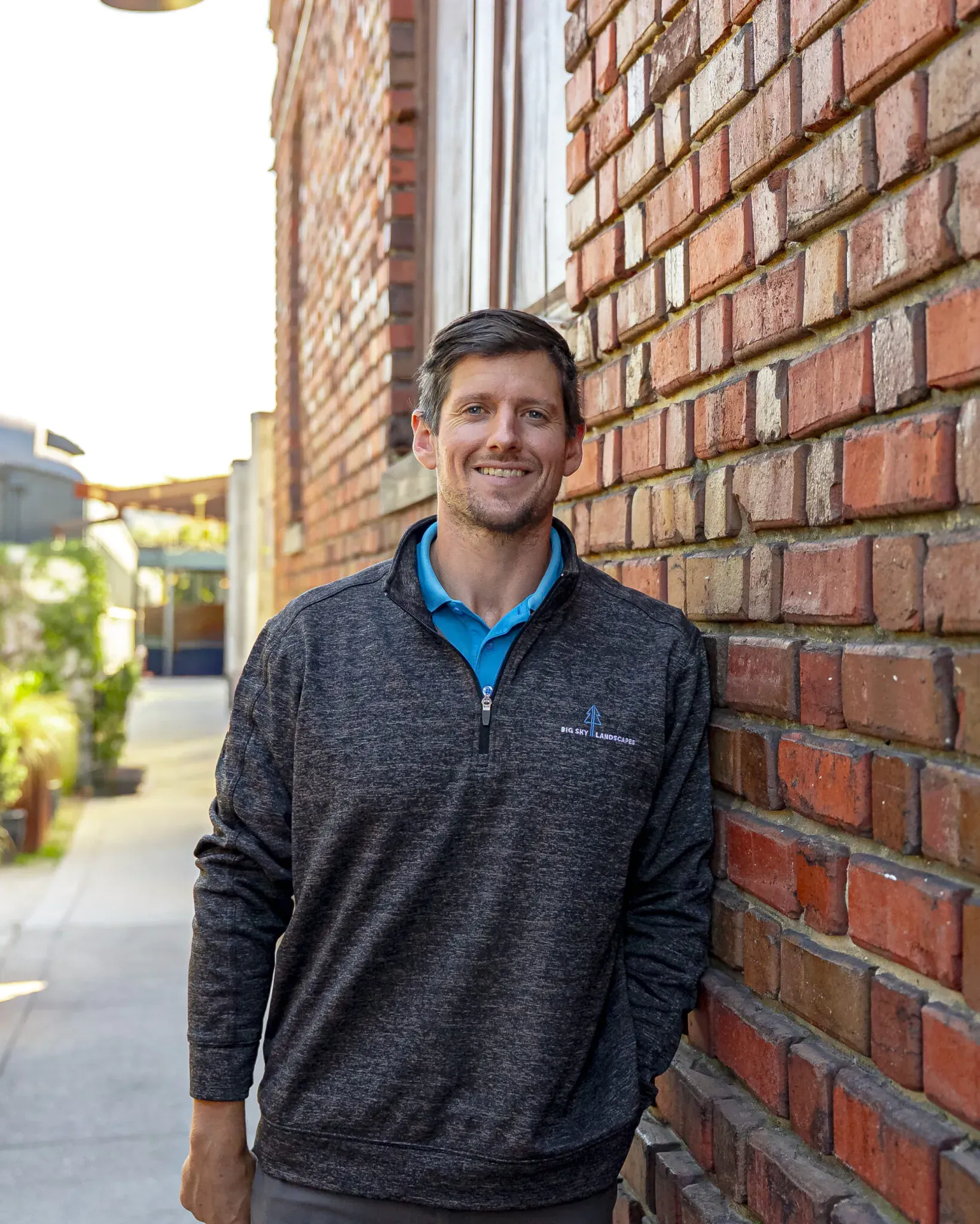 Smiling person in a dark sweater leaning against a brick wall outdoors.