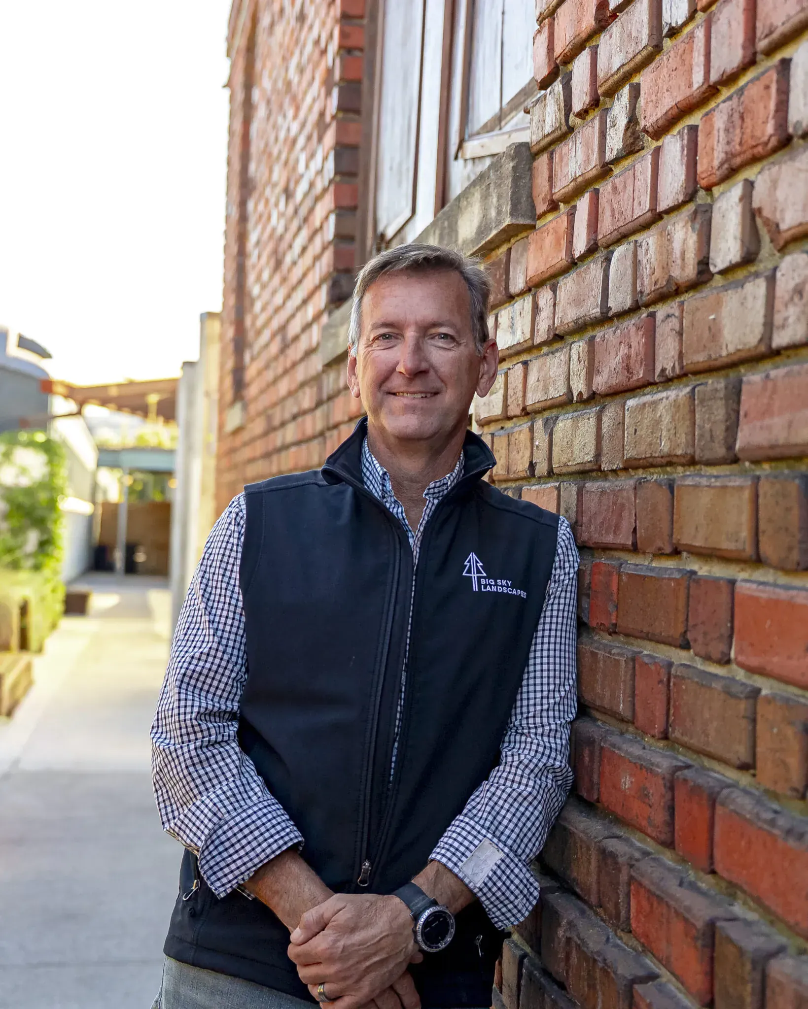 Person leaning against a brick wall outdoors, wearing a dark vest over a checkered shirt and smiling.