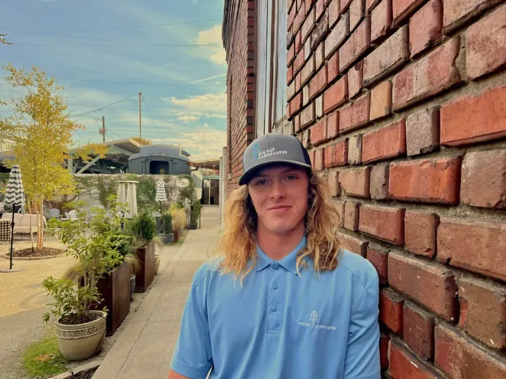 Person in a blue shirt standing by a brick wall on a sunny walkway with potted plants and trees.