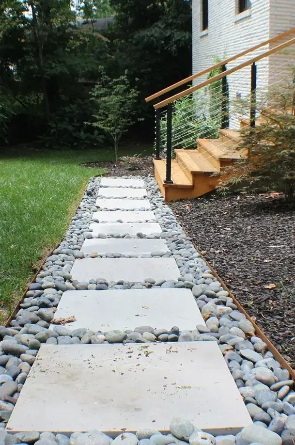 Stepping-stone walkway beside a house, bordered by river rocks, leading past wooden stairs and a grassy yard