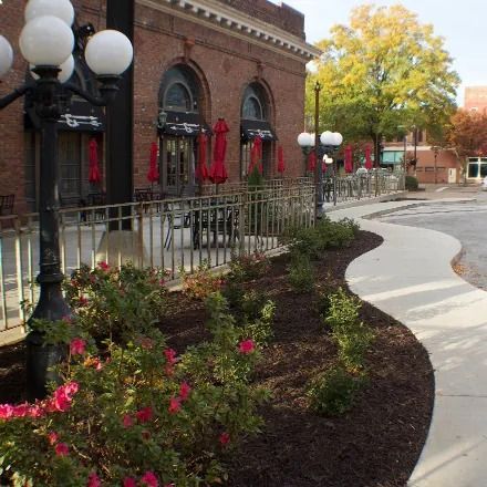 Exterior view of a brick building with a patio, red umbrellas, and a sidewalk lined with flower beds.