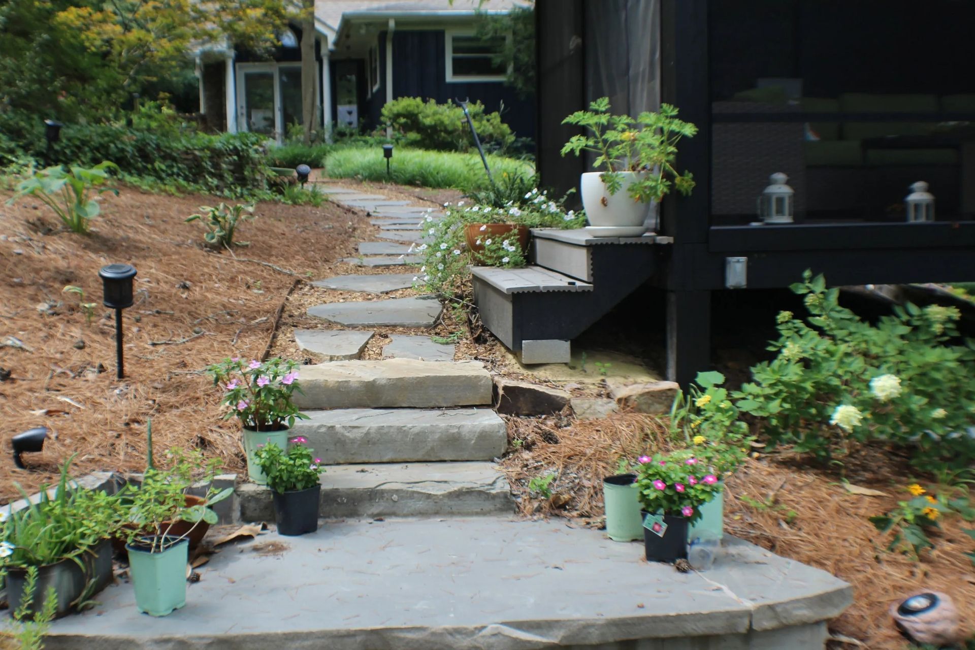 Stone pathway with steps leading up to a black deck, surrounded by greenery and potted plants.