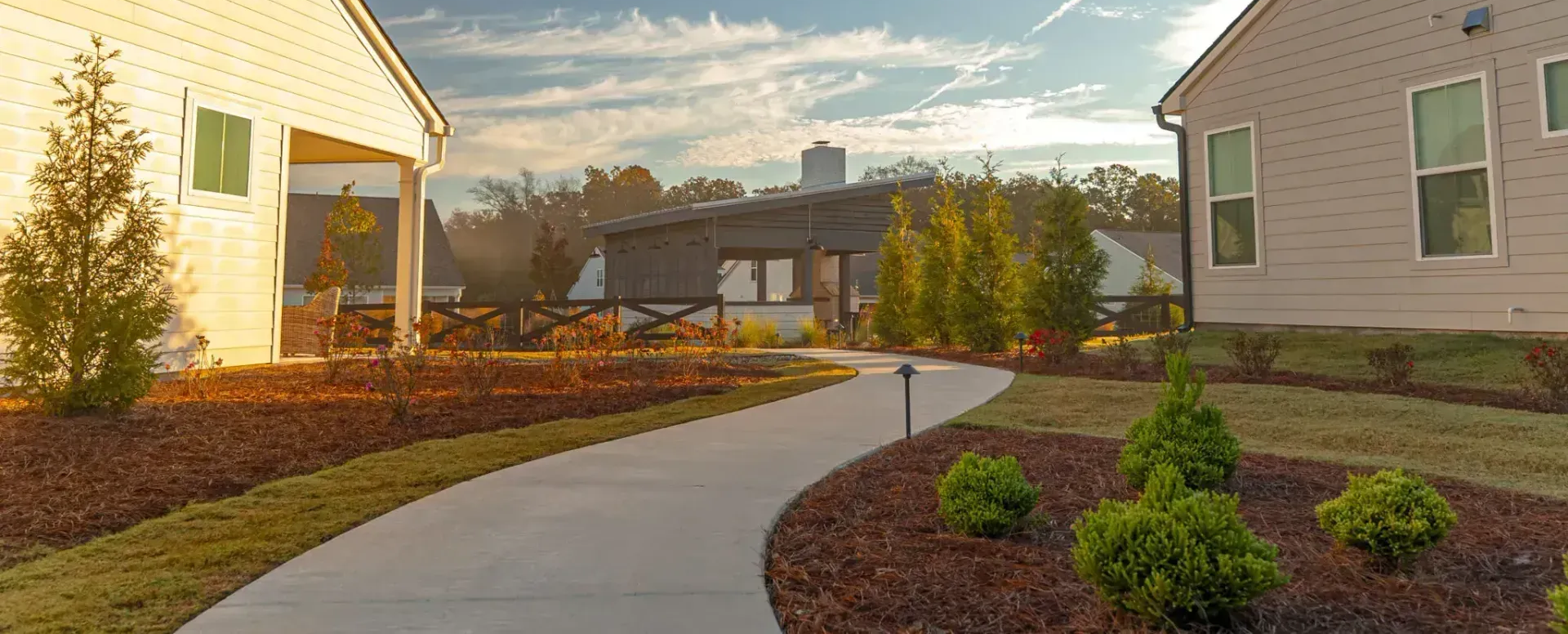Curving concrete walkway between landscaped yards, leading to a modern building under a cloudy sky.