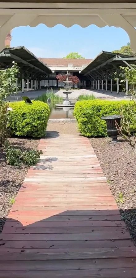A wooden path leads to a fountain and a building, flanked by green hedges and covered walkways.