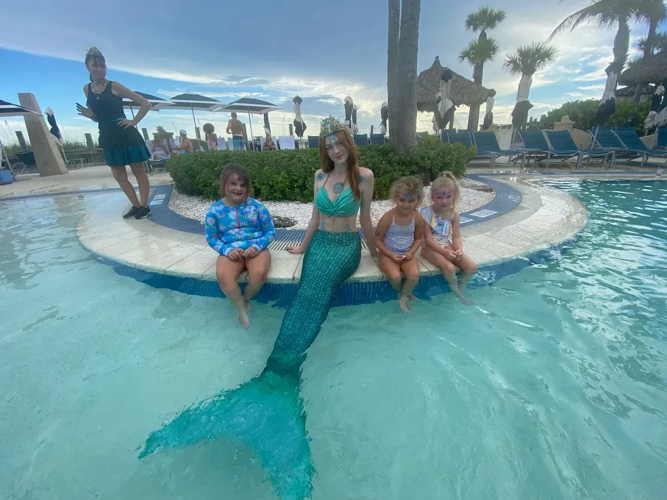 Children posing by a pool with a turquoise mermaid tail, in a sunny resort setting