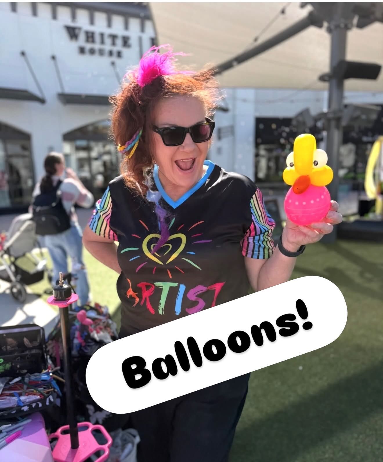 Woman with sunglasses holding pink and yellow balloon animal, wearing colorful shirt at outdoor event with “Balloons!” sign
