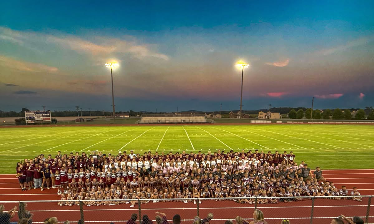 Football players huddle around a football, reaching hands together, blue sky backdrop.