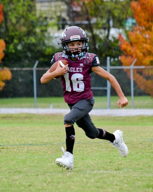Football players in action on a grassy field; one runs with the ball, pursued by two opponents.