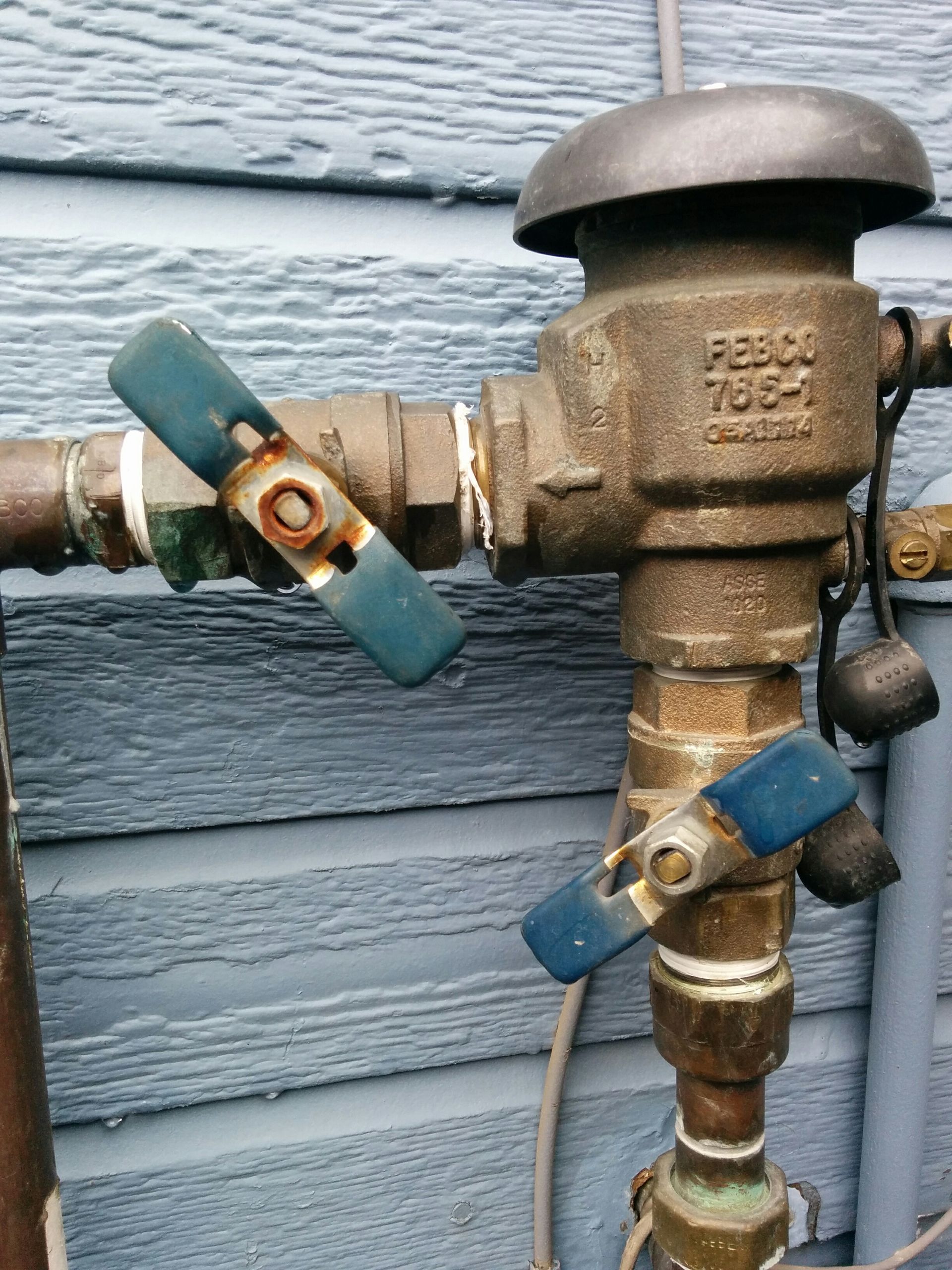 Bronze backflow preventer with blue handles, attached to pipes on a blue-sided building.