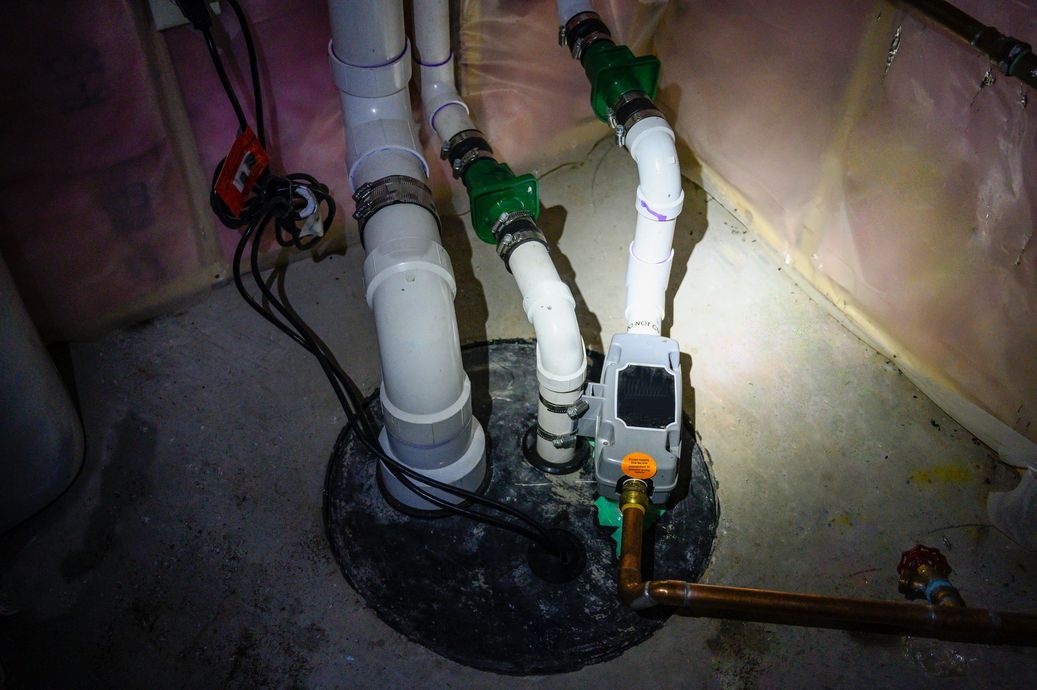 Sump pump system with white PVC pipes, green valves, and black basin in a basement.