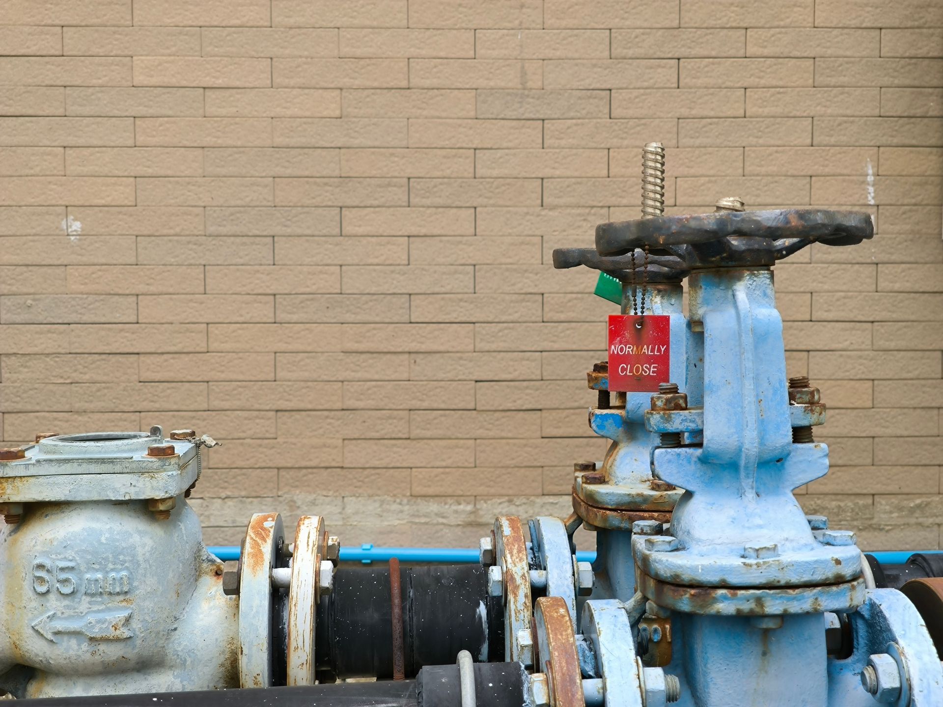 Blue water valves and pipes with tags against a tan brick wall background.