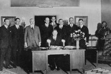 a black and white photo of a group of men standing around a desk .