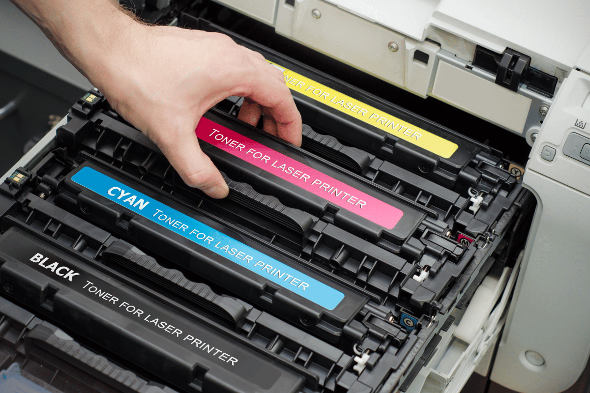 Hand inserting a magenta toner cartridge into a printer with cyan and black cartridges visible