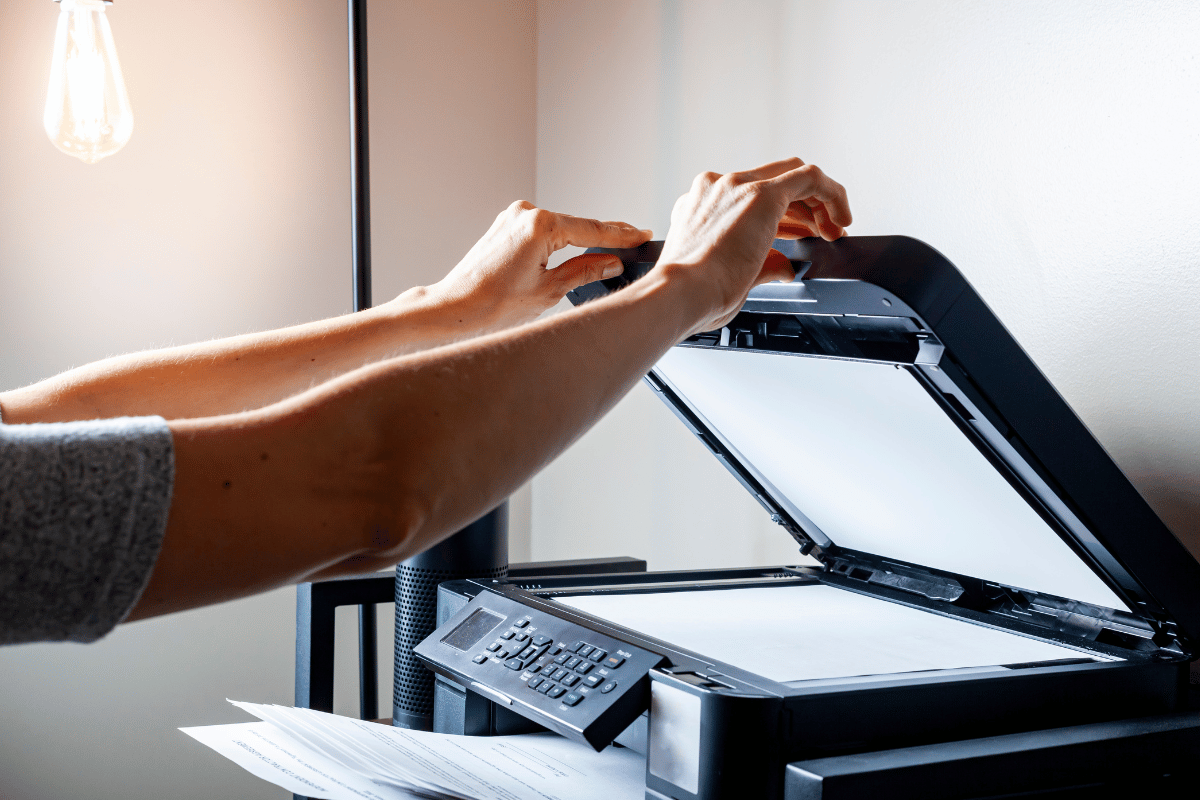 Person placing paper into an open flatbed scanner beside a desk lamp