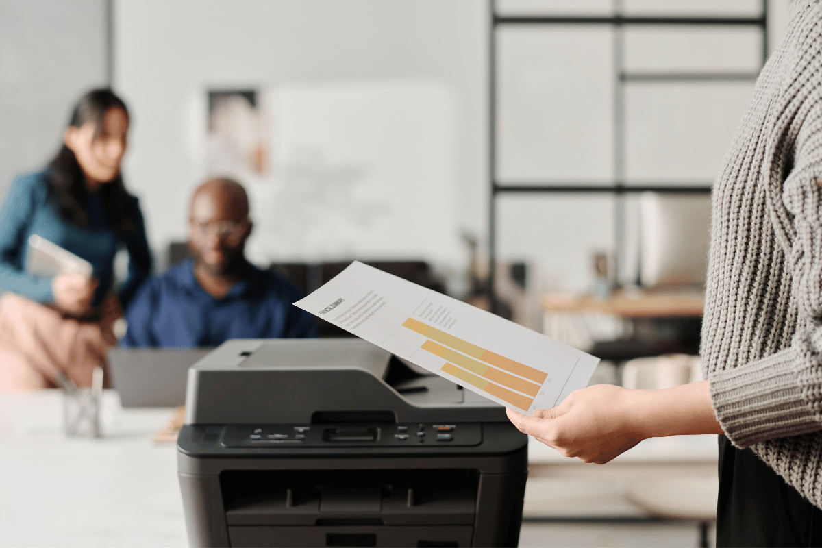 Office printer with a person holding a printed document, people working in the blurred background