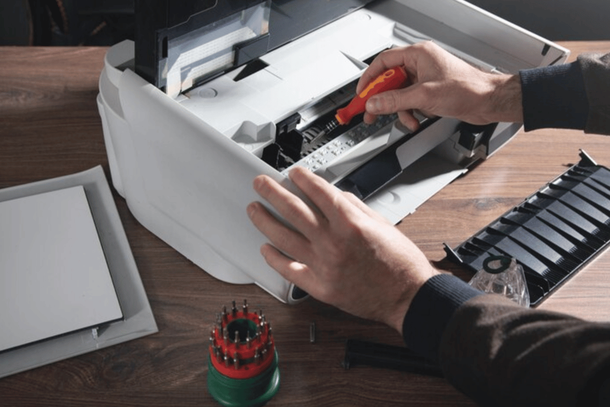 Hands adjusting a red ribbon in a white typewriter on a wooden desk with a screwdriver nearby