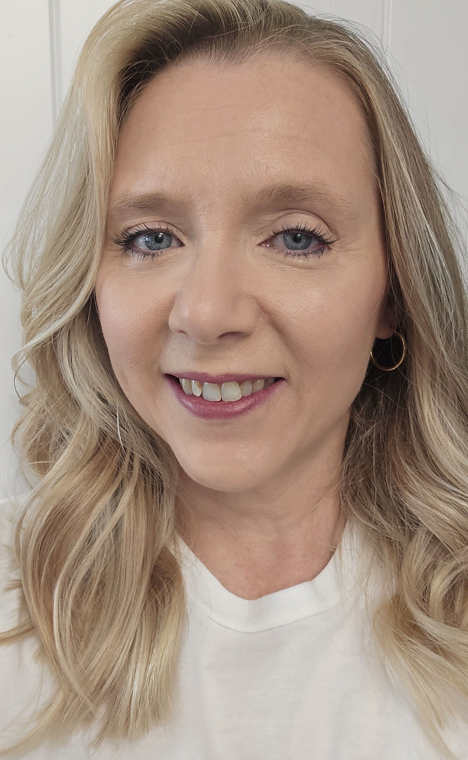 Blonde woman with blue eyes smiles, wearing a white shirt and hoop earrings.