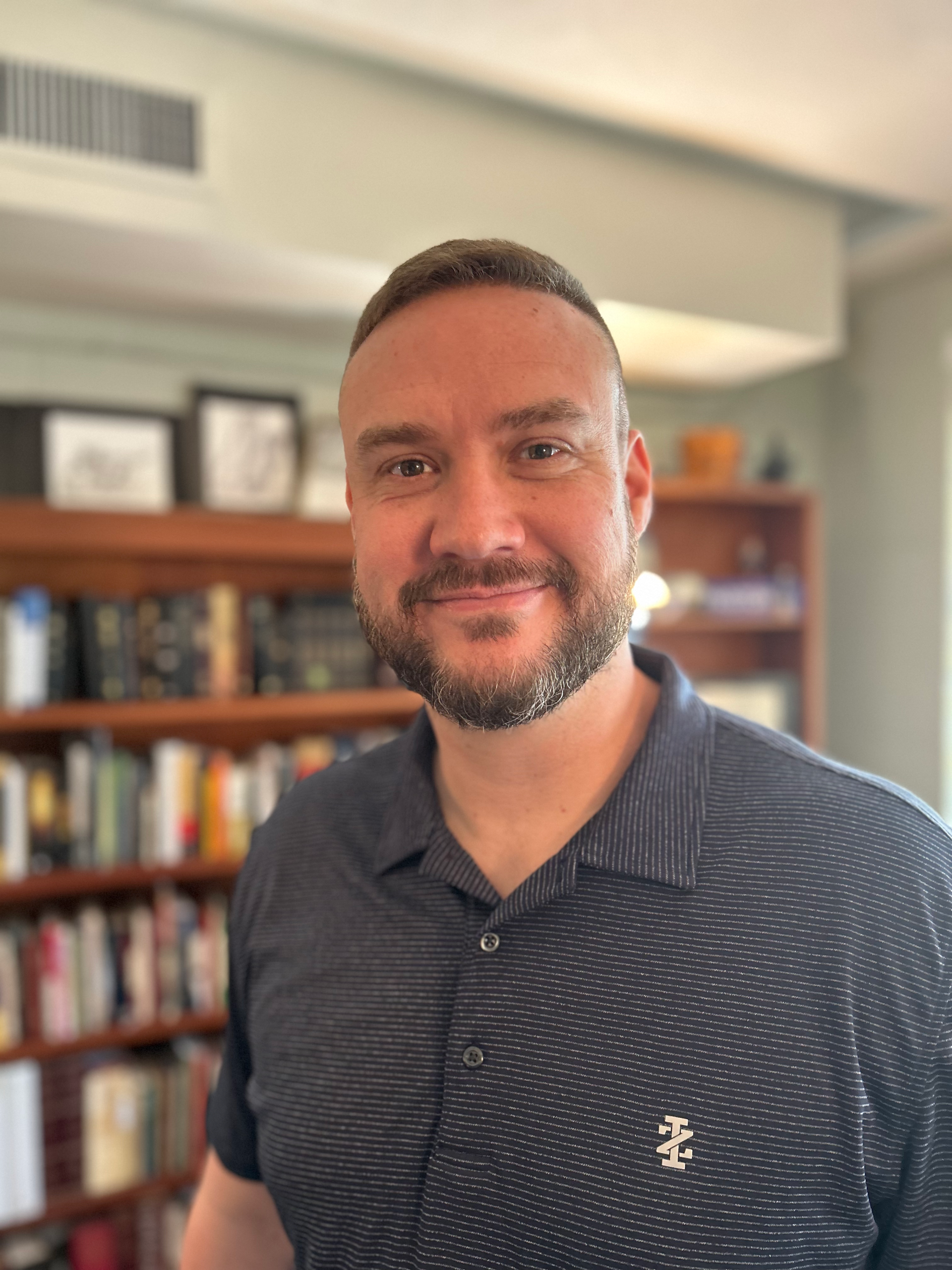 Man with a beard smiling, wearing a polo shirt, bookshelves in the background.