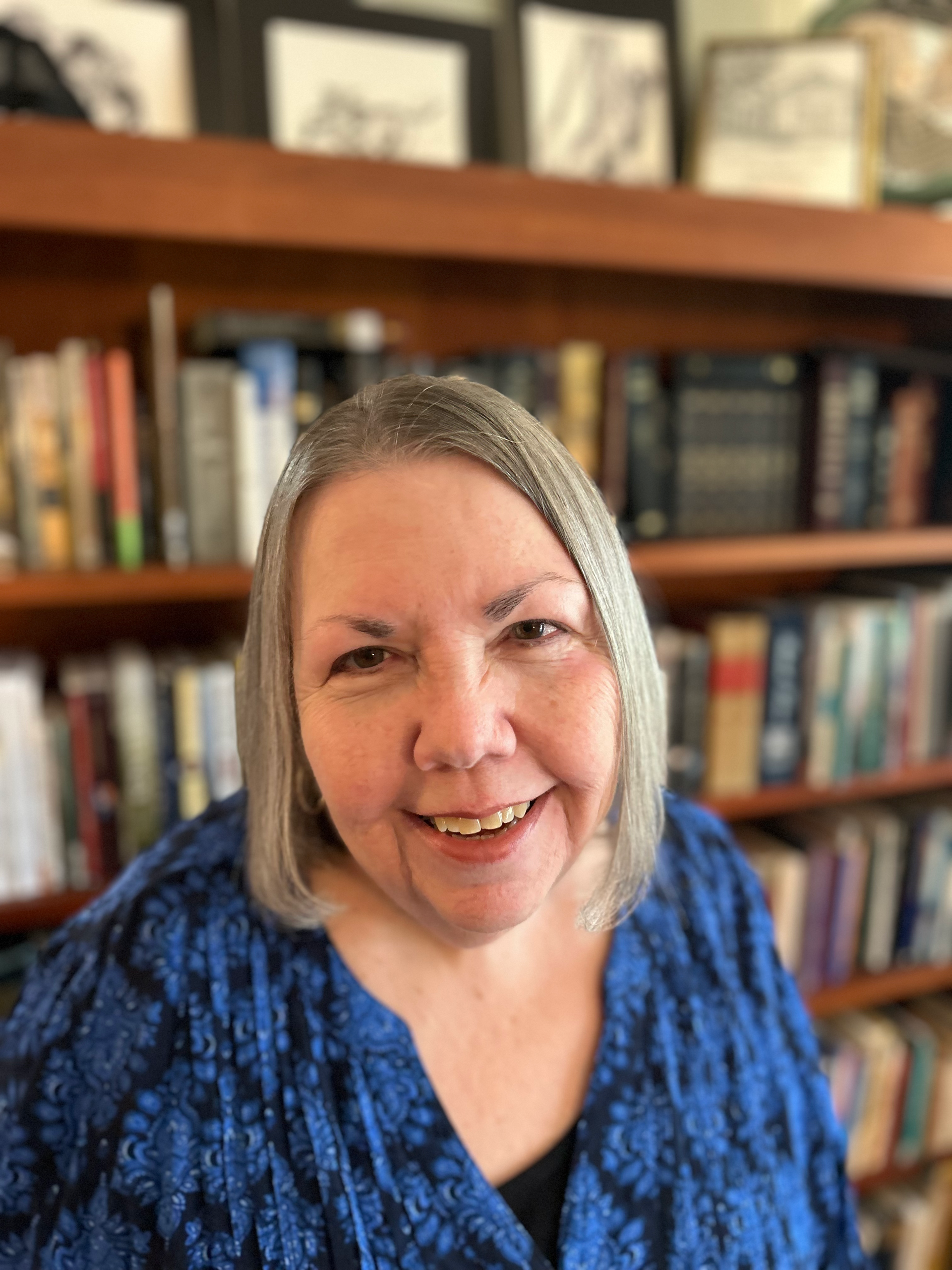 Woman with grey hair smiling, wearing a blue patterned shirt, in front of a bookshelf filled with books.