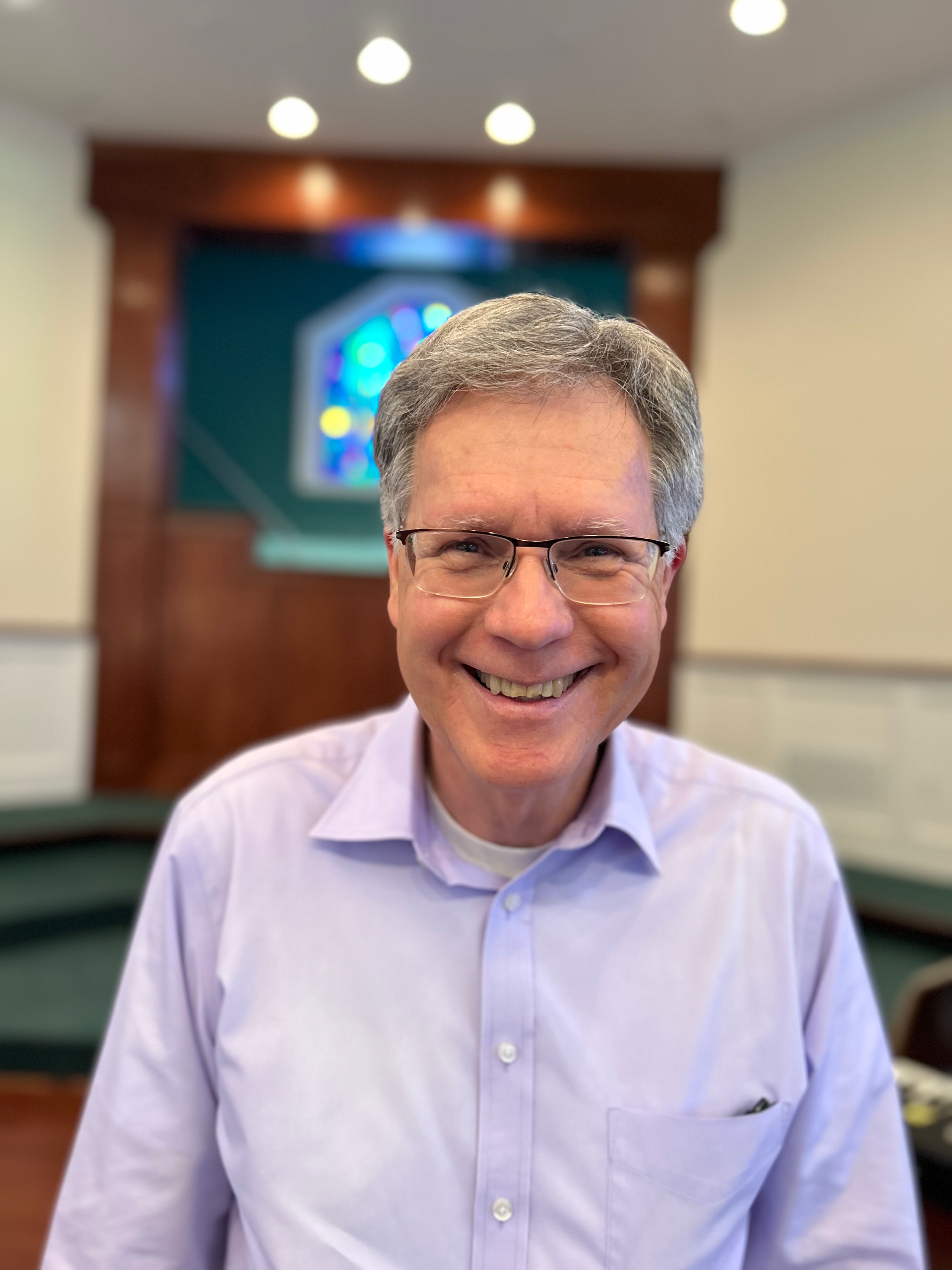 Man smiling, wearing glasses and a light purple shirt, standing in a church.