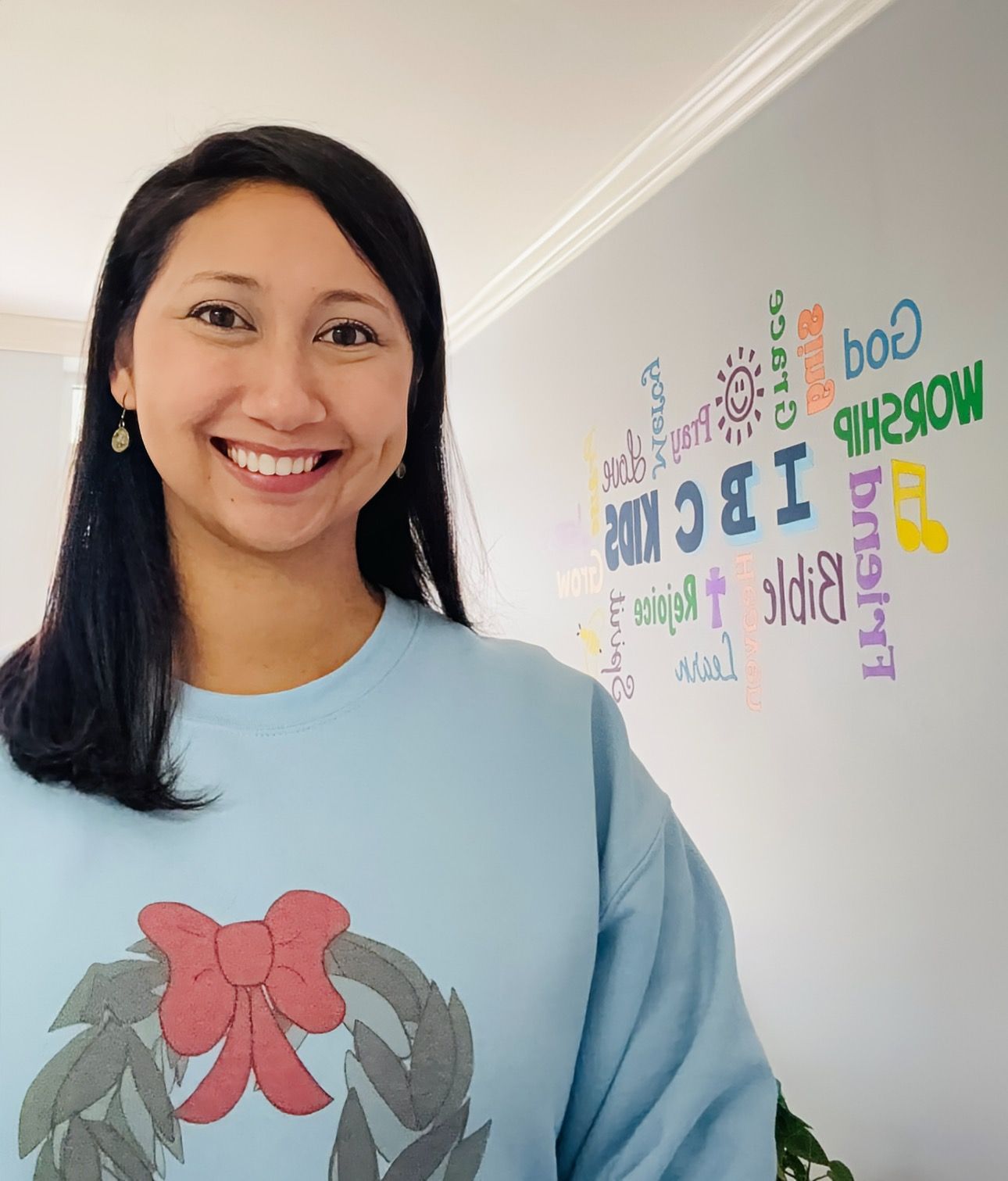 Woman smiles, wearing a light blue sweater with a wreath graphic. Wall has