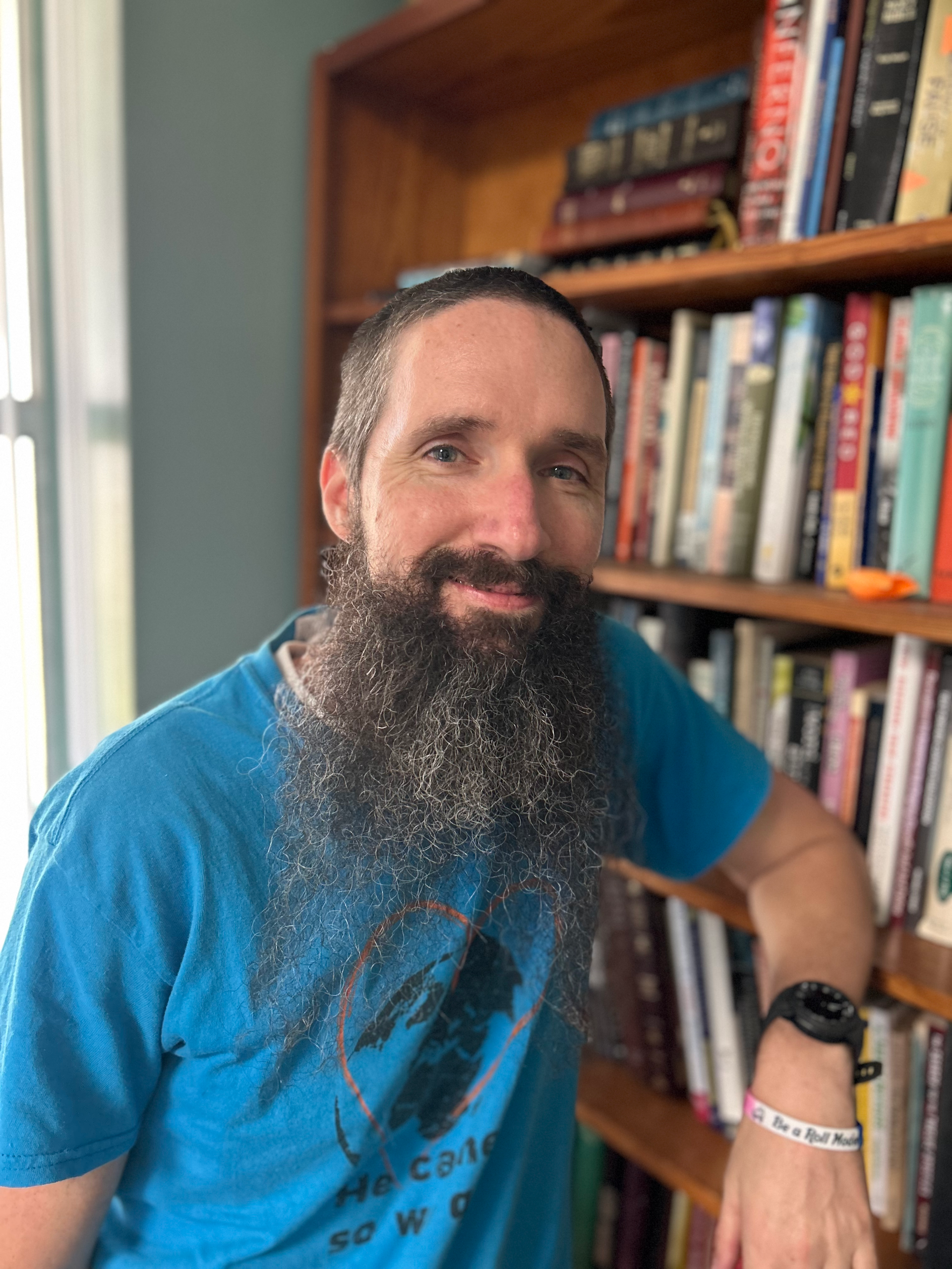 Man with a long beard in a blue shirt, leaning on a bookshelf in a room with a window.