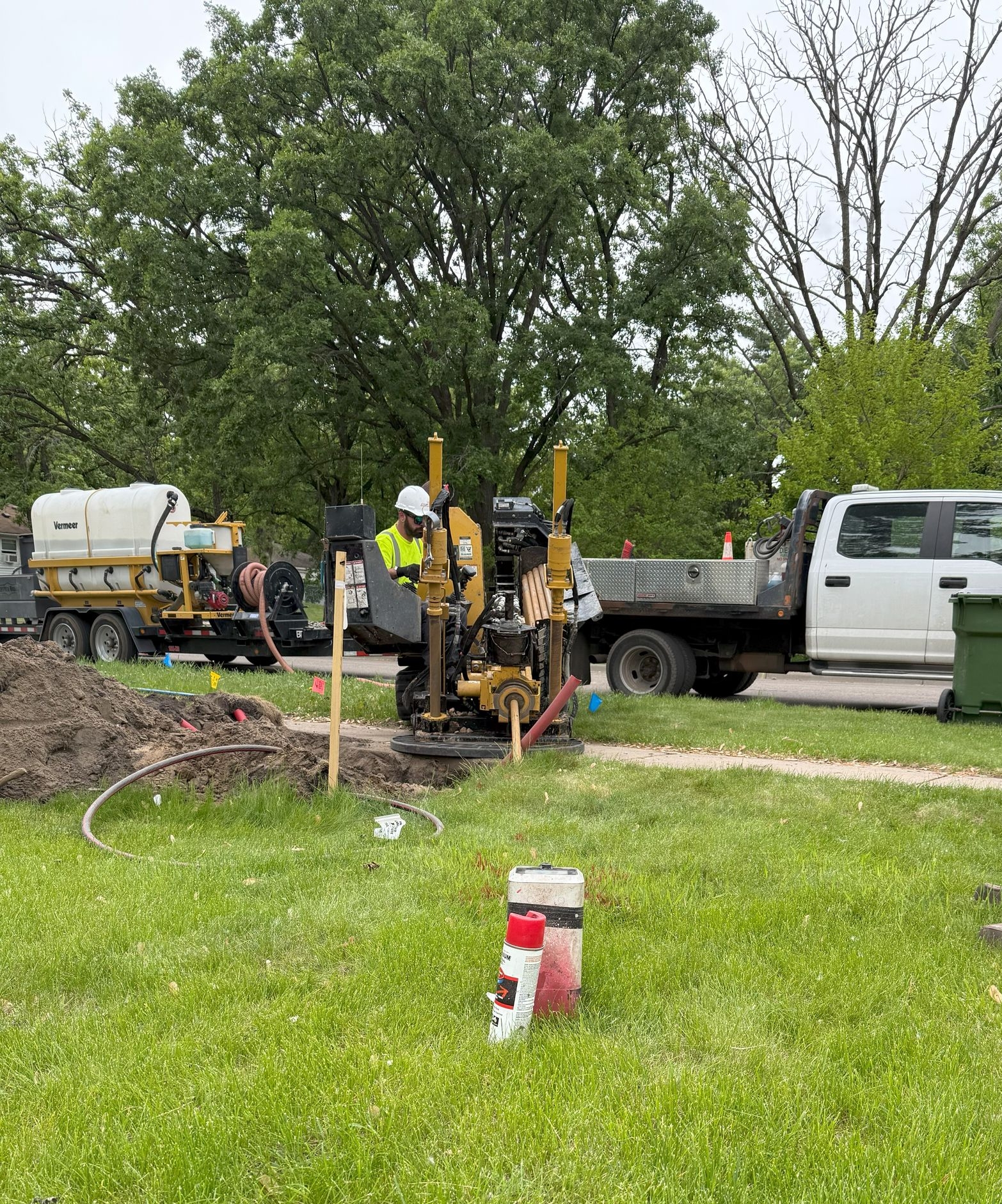 Construction worker operating drilling machine on a grassy area, with trucks and trees in the background.