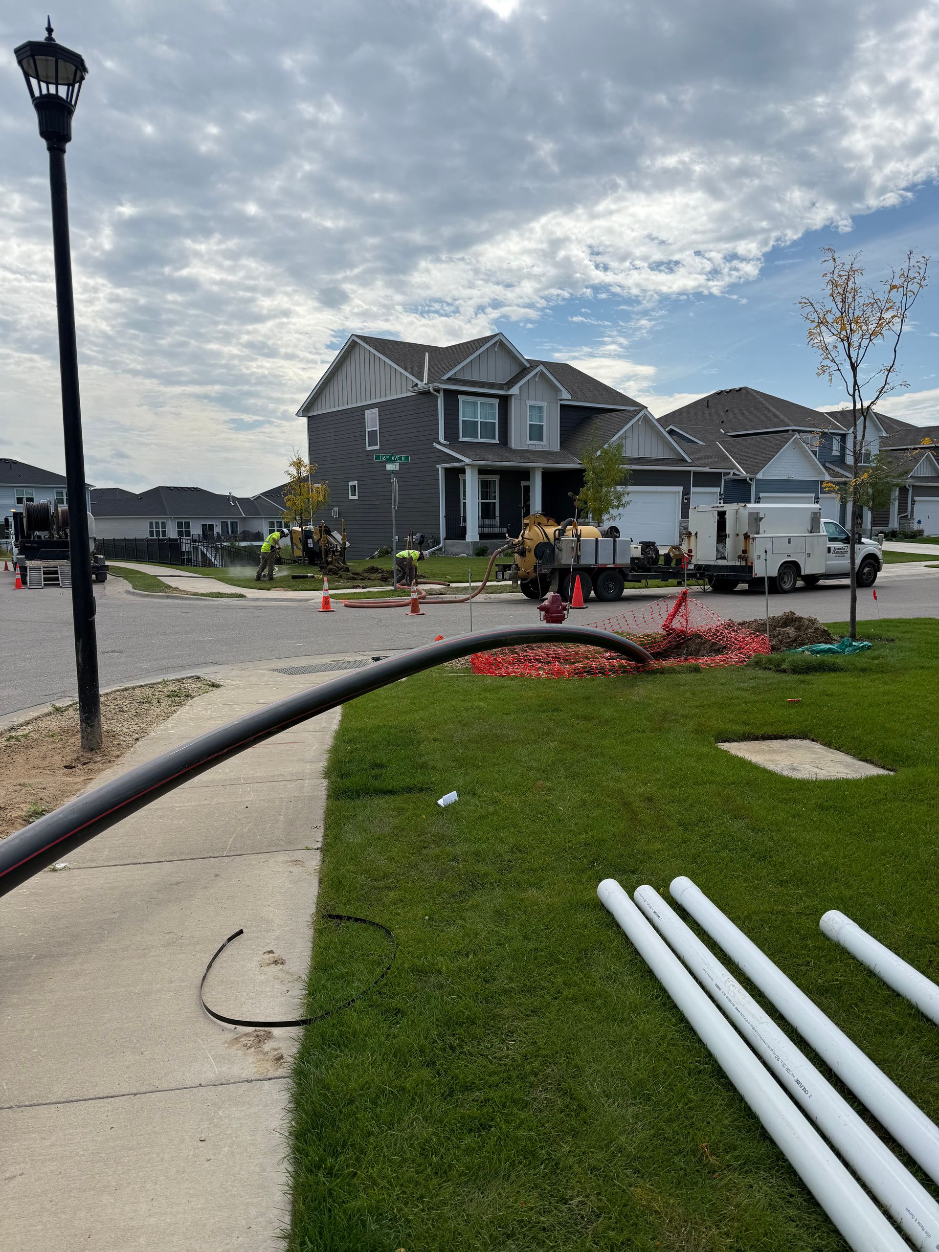 Construction site on residential street; black cable and white pipes on grass, workers near house.