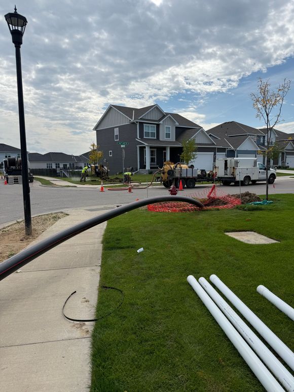 Construction site on residential street; black cable and white pipes on grass, workers near house.
