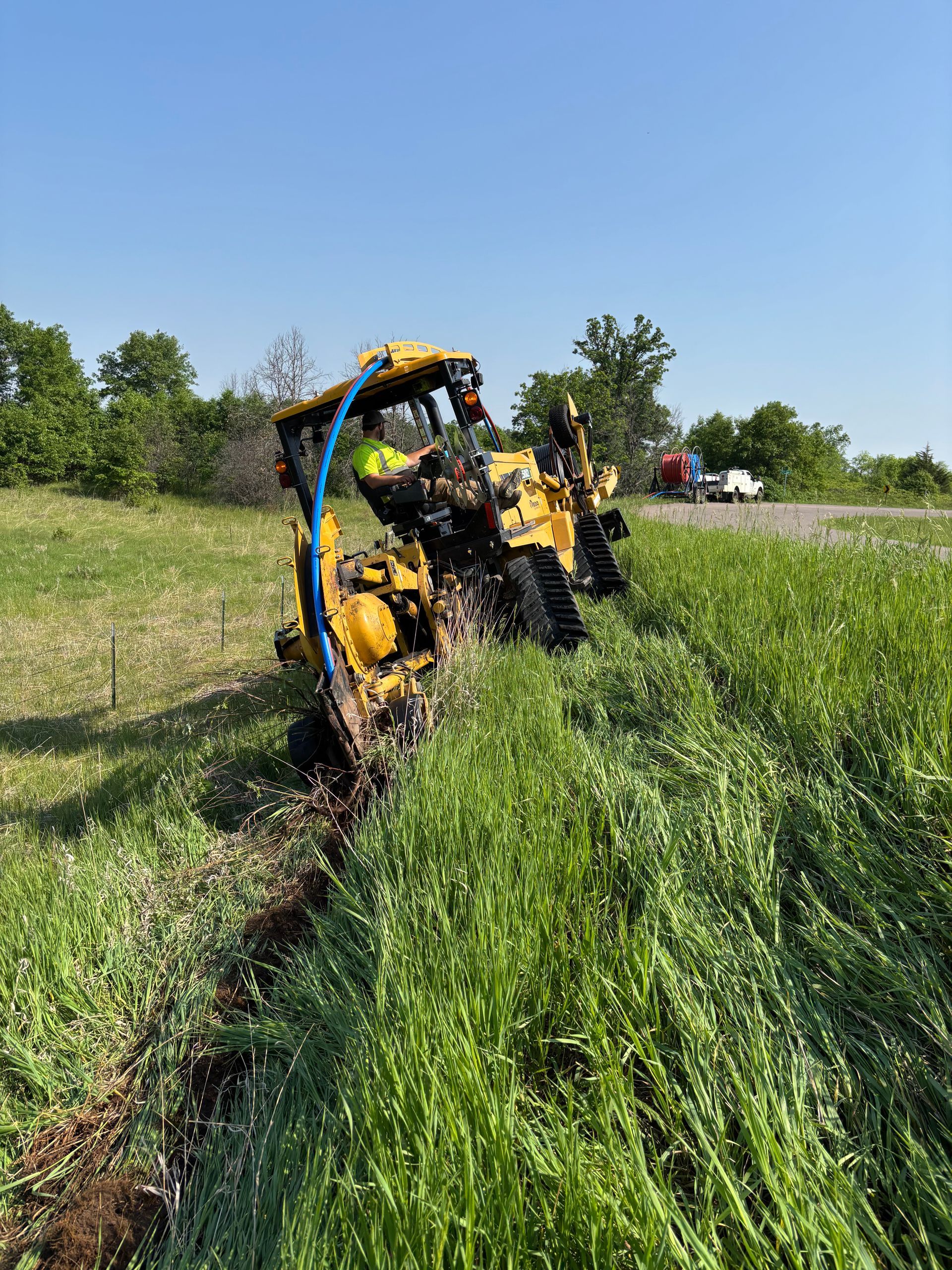 Yellow trencher digging in tall grass next to a road, blue sky overhead.