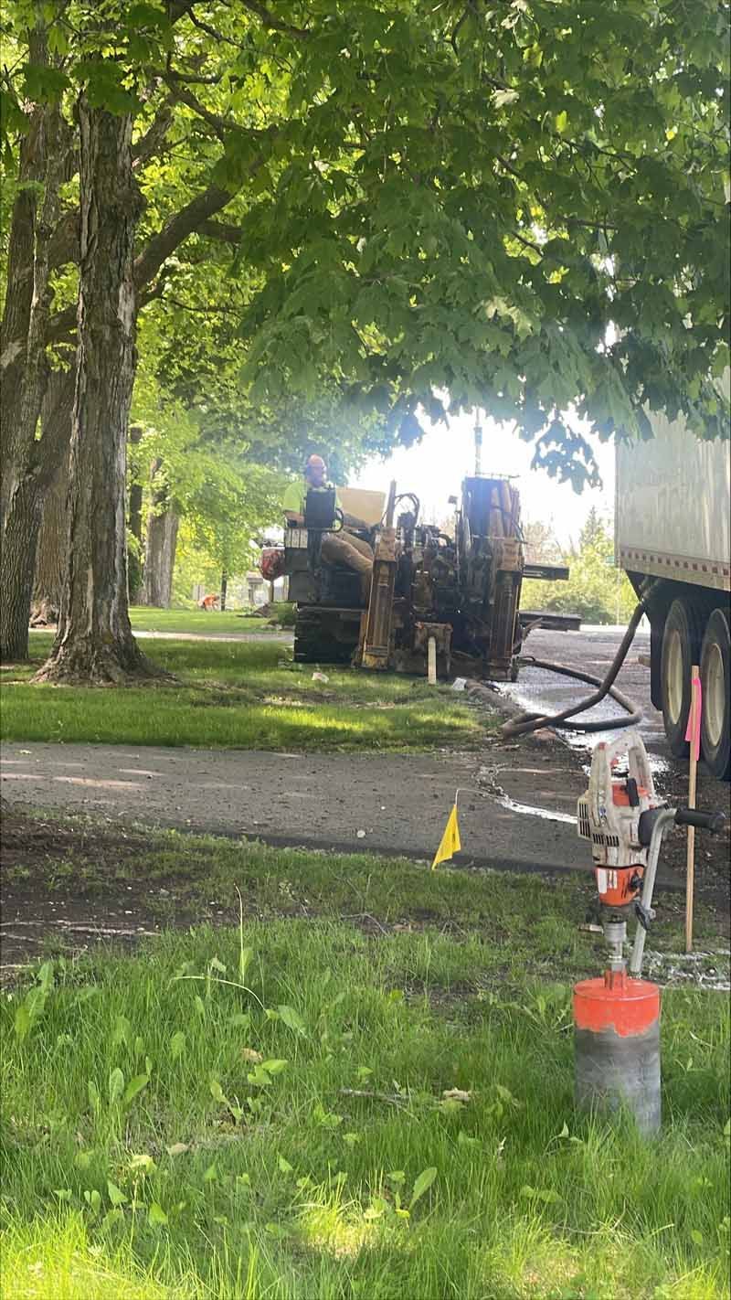 A utility crew operates heavy drilling machinery on a roadside near a neighborhood street with green trees.