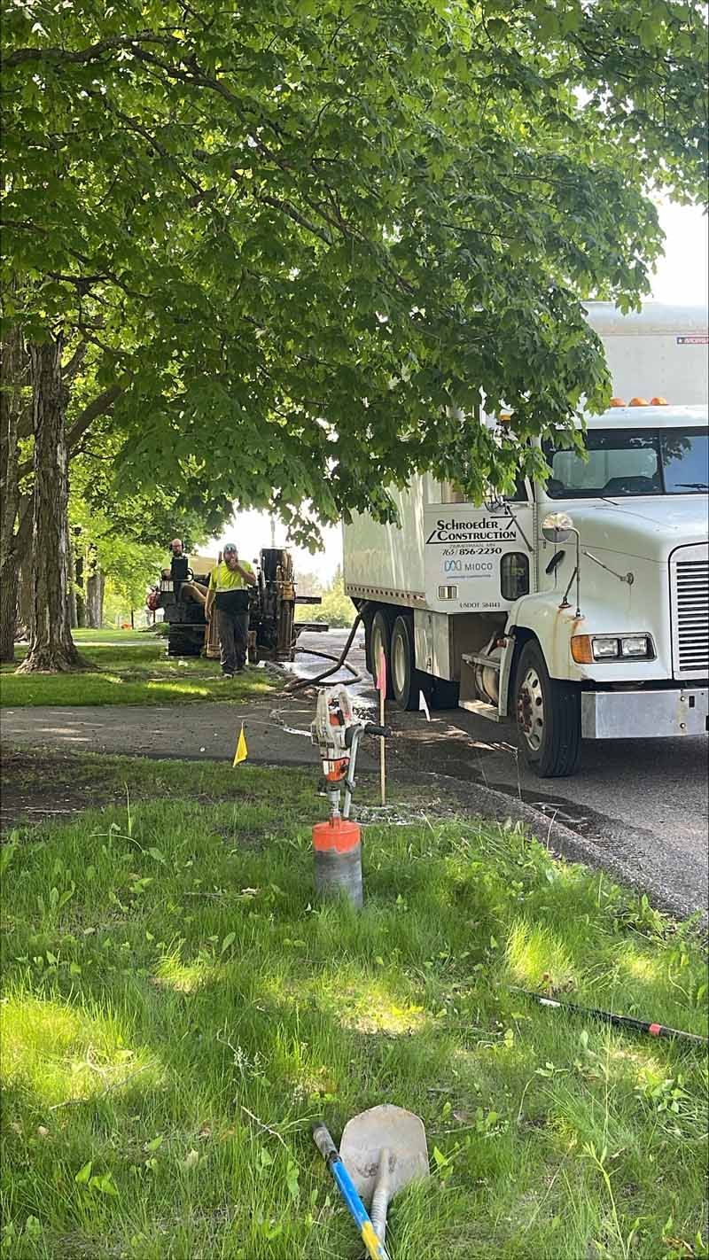 Well drilling operation next to a large truck and tree, with workers visible. A metal pipe protrudes from the ground.