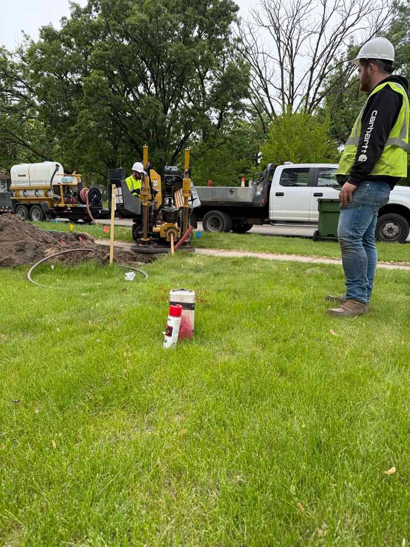 Man in safety vest observing drilling equipment on a grassy area near a truck.