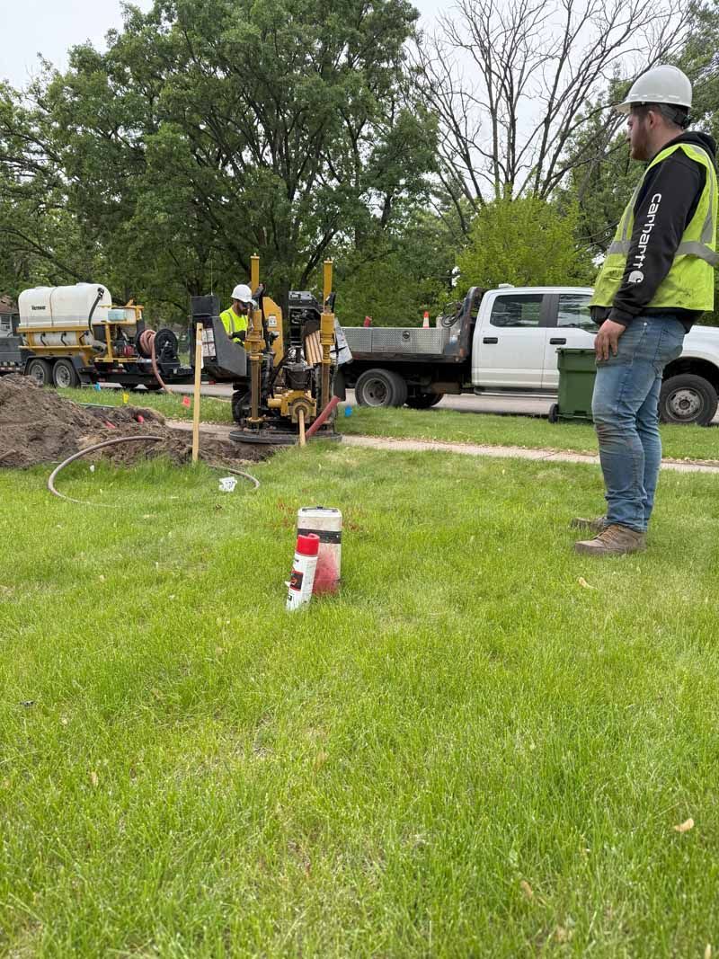 Workers operating machinery in a grassy area, a truck in background. A worker watches.