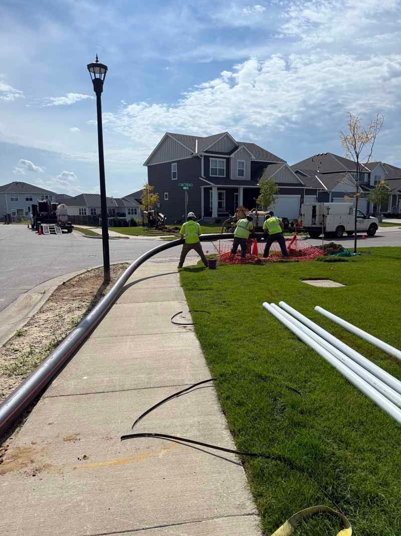 Construction workers installing pipes on a sidewalk near a residential neighborhood.
