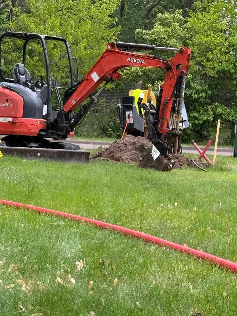 Red excavator digging in grass near a red utility line, next to a road and greenery.