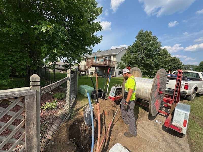 Two workers near a utility hole, fiber optic cable reel, truck, trees, and residential buildings.