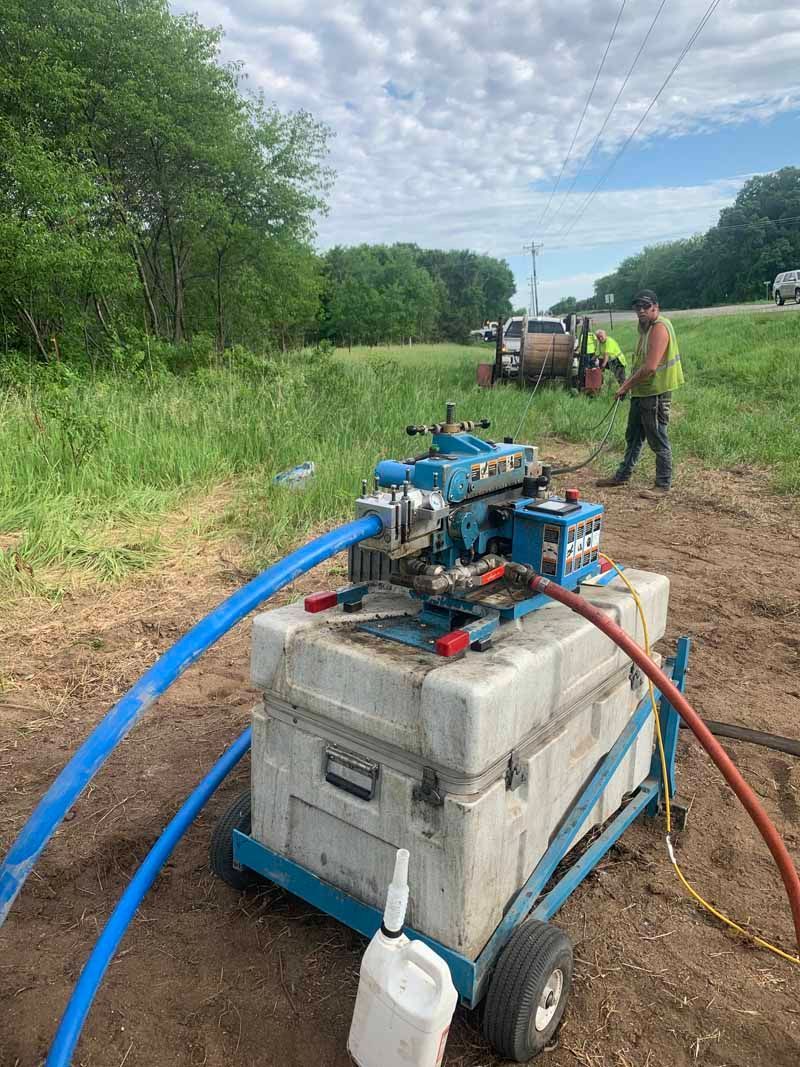 Blue machine with hoses on wheels, worker in vest nearby, roadside setting.