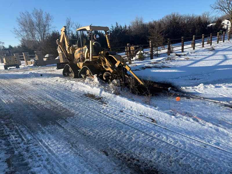 Backhoe clears snow from a road on a sunny day. Brown equipment, white snow, and a fence are visible.