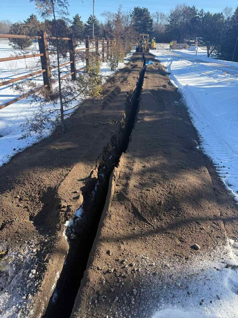 Trench dug in snow-covered ground alongside a wooden fence; a yellow backhoe is visible in the distance.