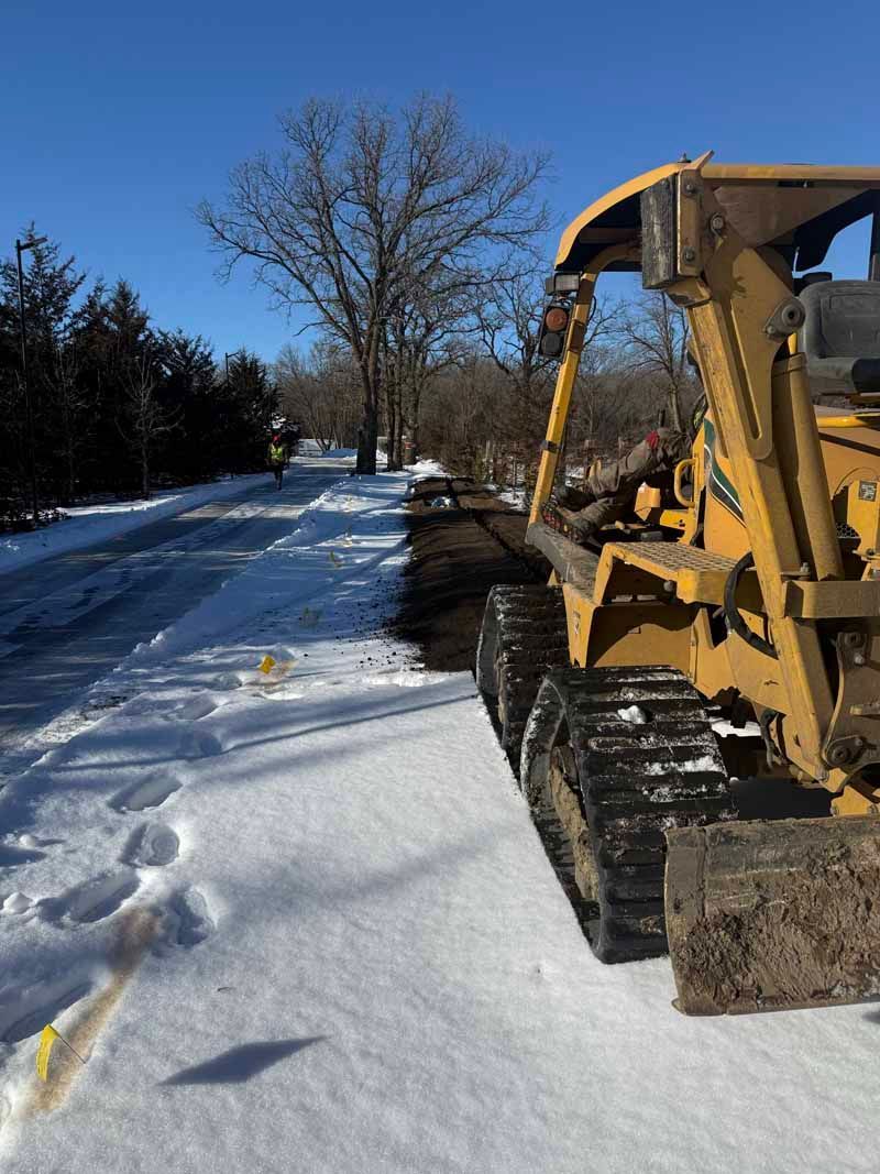 Yellow track skid steer next to a snow-covered road; a person in yellow jacket walks ahead on the road.