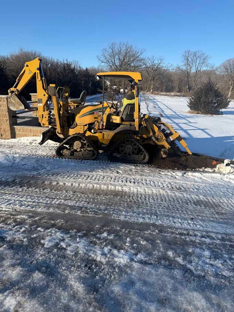 Yellow backhoe digging in snowy area, next to low wall.