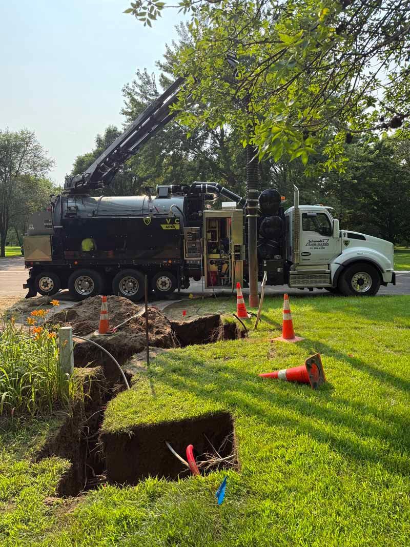 A large truck with an extended arm excavates a trench in a grassy yard, with traffic cones and visible utility lines.