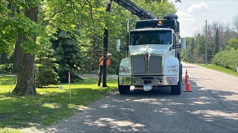 A utility truck with boom is next to a worker. Roadside with green trees.